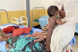 حوا تحمل ابنتها أوبا بينما يتمدد طفلها عبد الوالي مريضًا على السرير.