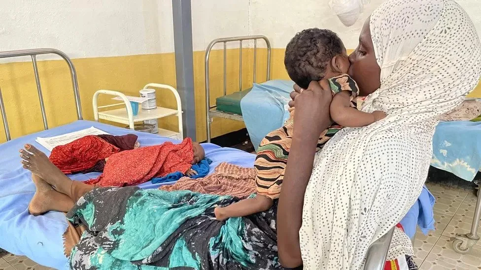 حوا تحمل ابنتها أوبا بينما يتمدد طفلها عبد الوالي مريضًا على السرير.